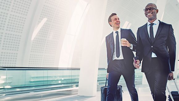 Two smiling businessman in suits talking together while walking with their baggage down a corridor in a bright modern airport